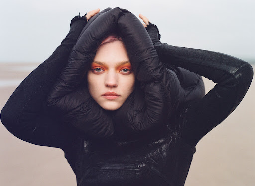 Close-up of a fair-skinned woman with pink hair and bright orange eyeshadow wearing a dark puffer jacket and standing on a beach.
