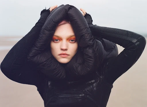 Close-up of a fair-skinned woman with pink hair and bright orange eyeshadow wearing a dark puffer jacket and standing on a beach.