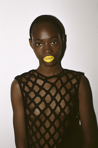 Close-up portrait of a dark-skinned woman with yellow lipstick wearing a black crochet top against a plain white background, beauty shot.