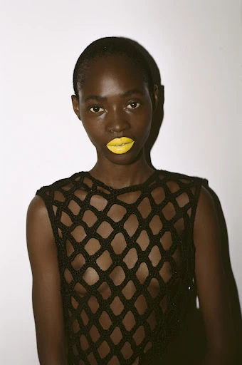 Close-up portrait of a dark-skinned woman with yellow lipstick wearing a black crochet top against a plain white background, beauty shot.