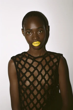 Close-up portrait of a dark-skinned woman with yellow lipstick wearing a black crochet top against a plain white background, beauty shot.
