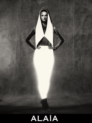Black and white portrait of a woman with hands on hips wearing a white dress and head covering in a studio setting.
