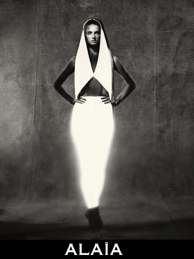 Black and white portrait of a woman with hands on hips wearing a white dress and head covering in a studio setting.
