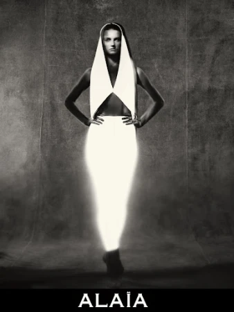 Black and white portrait of a woman with hands on hips wearing a white dress and head covering in a studio setting.