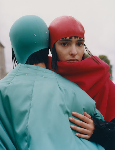 Two models pose in an outdoor setting with muted colors; one wears a red hat and the other wears a turquoise head covering and jacket.