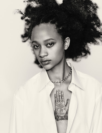 Black-and-white portrait of a woman with voluminous dark hair wearing a white collared shirt and a hand tattoo on her chest.