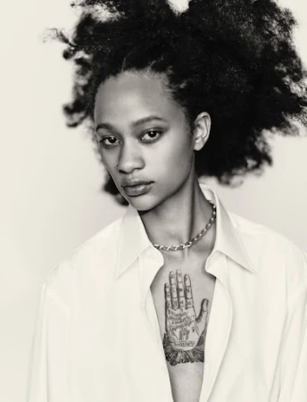 Black-and-white portrait of a woman with voluminous dark hair wearing a white collared shirt and a hand tattoo on her chest.