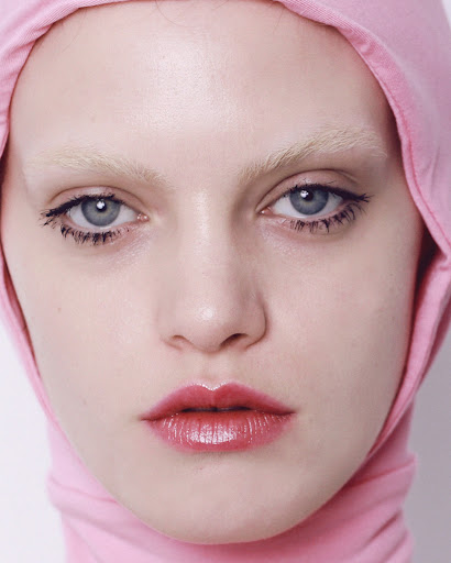 Close-up of a fair-skinned young woman with bleached eyebrows and pink lipstick, wearing a light pink hooded garment.