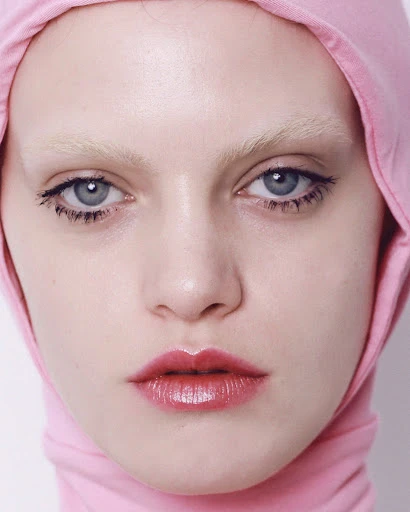 Close-up of a fair-skinned young woman with bleached eyebrows and pink lipstick, wearing a light pink hooded garment.