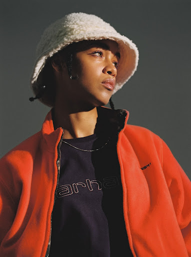 Portrait of a young adult woman with dark hair wearing an orange fleece jacket and a fuzzy bucket hat looking off to the side.