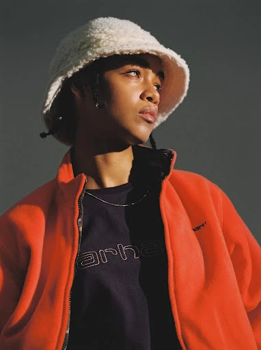 Portrait of a young adult woman with dark hair wearing an orange fleece jacket and a fuzzy bucket hat looking off to the side.