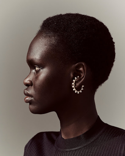 Profile of a dark-skinned adult female wearing a black ribbed top and pearl hoop earring against a neutral grey background in studio lighting.
