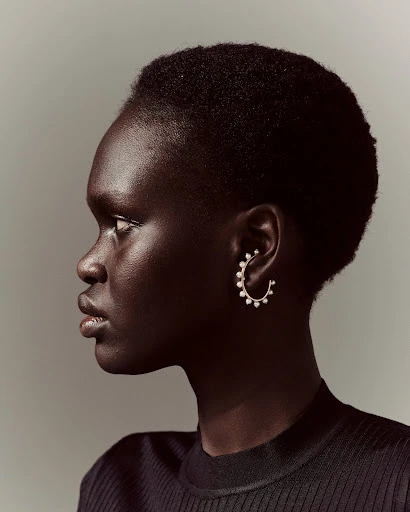 Profile of a dark-skinned adult female wearing a black ribbed top and pearl hoop earring against a neutral grey background in studio lighting.
