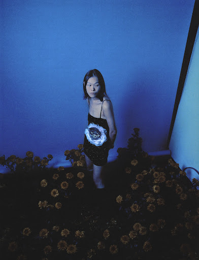 Model standing among dark flowers wearing a black dress under cool blue lighting by makeup artist Yvane Rocher