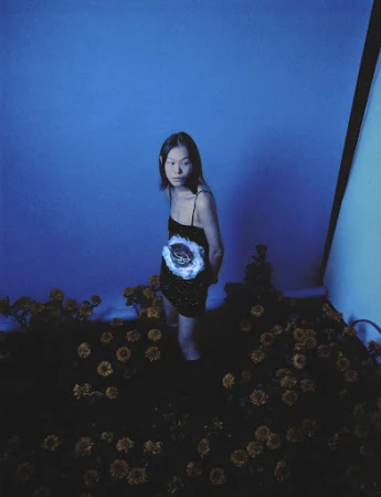 Model standing among dark flowers wearing a black dress under cool blue lighting by makeup artist Yvane Rocher