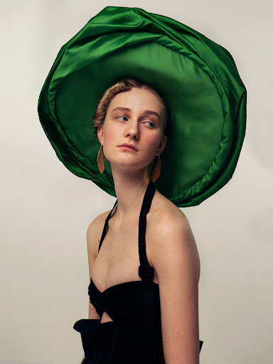 Model wearing a large green satin hat and black halter top with gold earrings against a plain background