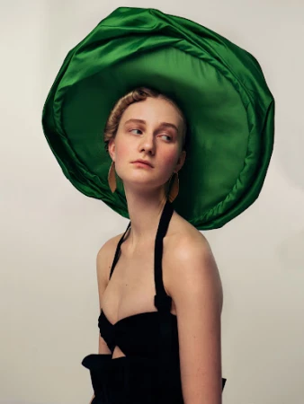 Model wearing a large green satin hat and black halter top with gold earrings against a plain background