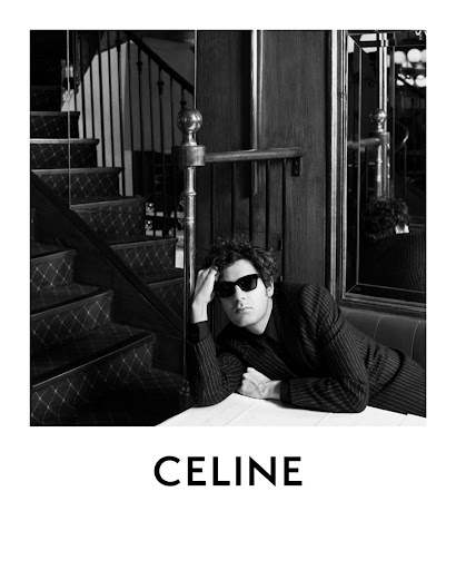 Black and white shot of a man in sunglasses and a pinstripe suit leaning on a table, with a staircase and the CELINE logo visible.