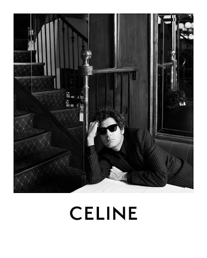 Black and white shot of a man in sunglasses and a pinstripe suit leaning on a table, with a staircase and the CELINE logo visible.