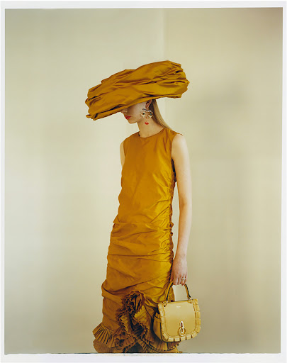 An adult female fashion model wearing a large yellow hat and yellow dress, holding a purse, is posing in a photo studio.