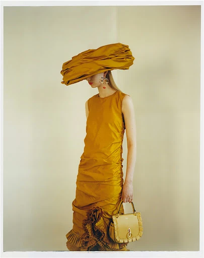 An adult female fashion model wearing a large yellow hat and yellow dress, holding a purse, is posing in a photo studio.