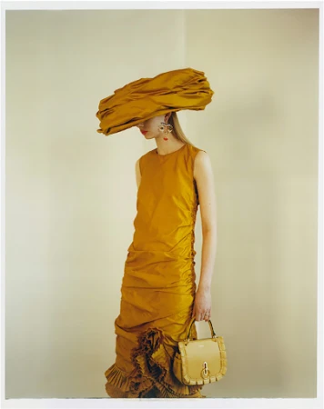 An adult female fashion model wearing a large yellow hat and yellow dress, holding a purse, is posing in a photo studio.