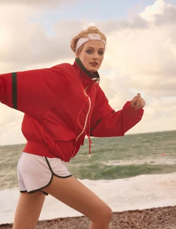 An adult female is shown jogging on a beach. She wears a red tracksuit jacket, shorts with trim, and a headband. The background shows the ocean.