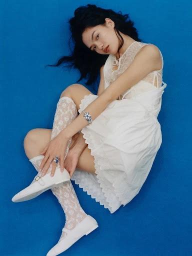 Young woman poses in a white lace dress with blue jewellery against a blue background, wearing white socks and white Mary Jane shoes.