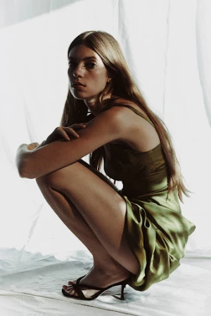 Model with long brown hair wearing a green silk dress crouching against a draped white fabric background