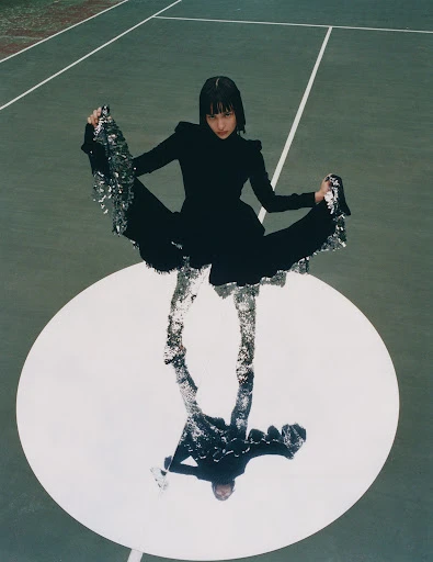 Woman in elegant black dress poses on a tennis court, her reflection visible on a circular mirror beneath her feet, in high-angle shot.