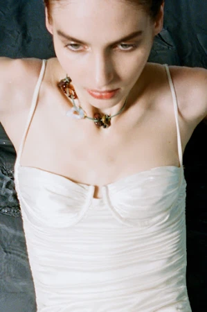 High-angle portrait of a woman wearing a white satin strapless top and a beaded necklace against a dark textured background.