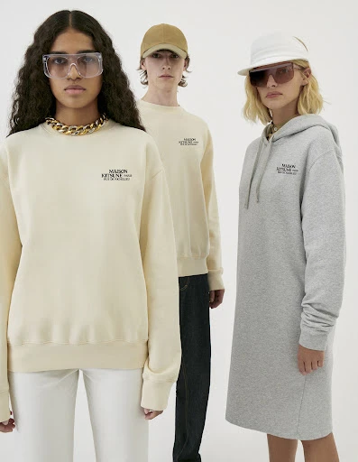 Three models showcase Maison Kitsuné apparel, featuring sweatshirts and a hoodie dress against a clean white backdrop in a studio shot.