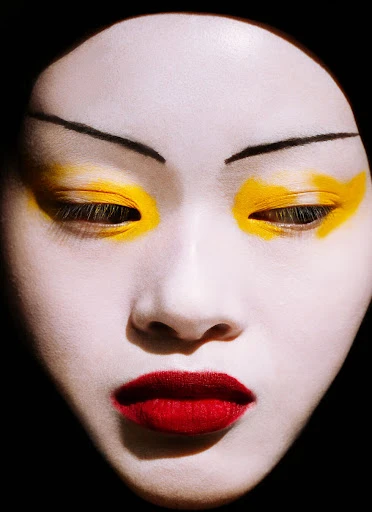 Close-up portrait of a person with white face paint, yellow eye makeup, and red lipstick against a dark background. Beauty concept.