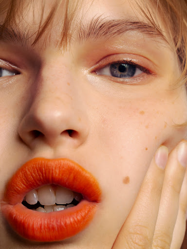 Close-up portrait of a fair-skinned adult female with orange lipstick, blue eyes, and light brown hair with hand near face. Beauty shot.