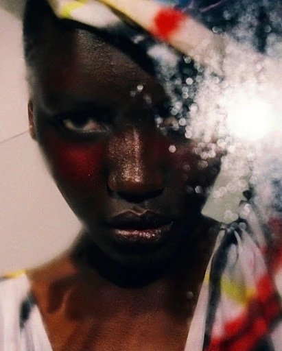Close-up portrait of a dark-skinned person with red face paint, a colorful head covering, and a blurred, bokeh-filled foreground.