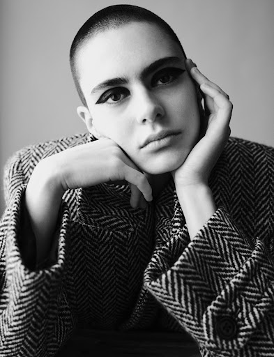 Black and white portrait of a woman with a shaved head and dramatic winged eyeliner, wearing a herringbone patterned jacket indoors.