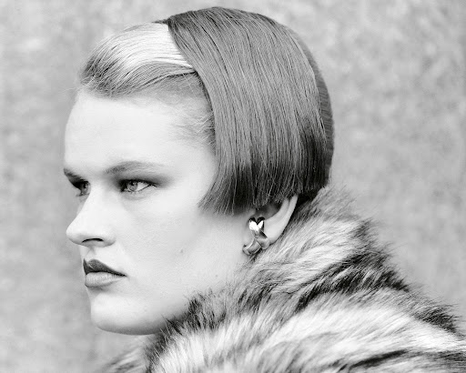 Black and white headshot of a fashionable adult woman with short, dark hair and fair skin, wearing a faux fur coat and heart earrings.
