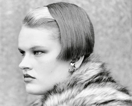 Black and white headshot of a fashionable adult woman with short, dark hair and fair skin, wearing a faux fur coat and heart earrings.