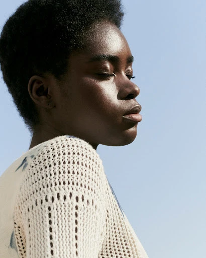 Close-up studio shot of a dark-skinned young adult woman with short hair against a light blue sky, wearing a white crocheted top.