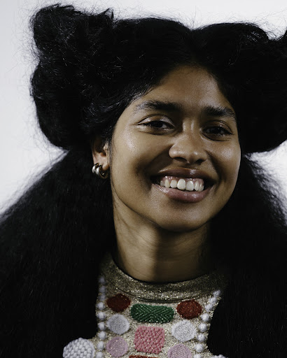 Portrait of a smiling East Asian woman with styled black hair, wearing a patterned beaded top against a plain white studio backdrop.