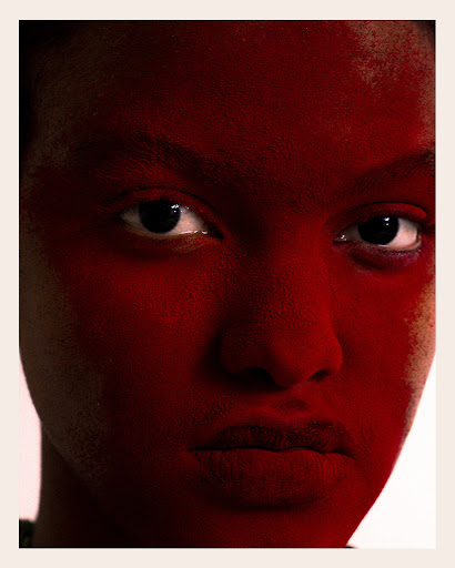 Close-up portrait of a young woman with dark hair and skin, her face painted in red tones, set against a soft, diffused light background.