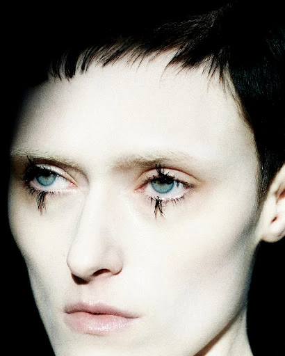 Close-up portrait of a pale-skinned person with dark, short hair and blue eyes featuring graphic black makeup, set against a dark background.