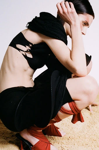 Woman crouches on sand in black bra top, shorts, and red wrap heels. She has dark hair, pale skin, and thin build. Studio shot.