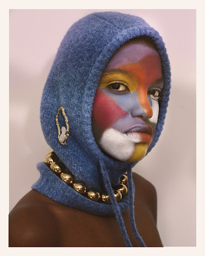 Portrait of a dark-skinned woman with colorful face paint wearing a blue knit balaclava, gold bead necklace and earring.