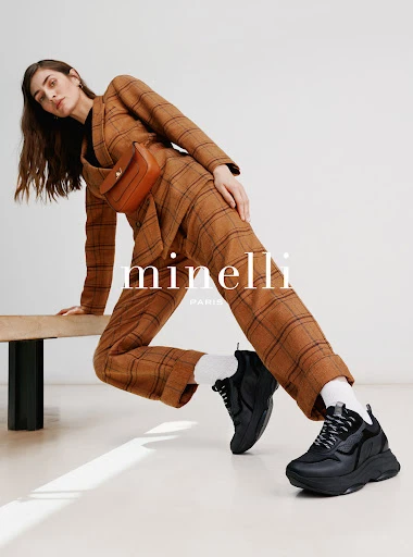 Fashion editorial shot of a model in a brown plaid suit and black sneakers, posing against a light grey background in a studio.