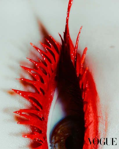 Macro beauty shot of a brown eye with dramatic, elongated bright red artistic eyelashes and makeup against a light grey background.