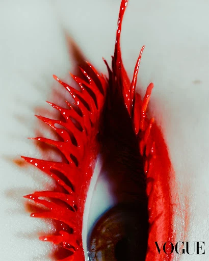 Macro beauty shot of a brown eye with dramatic, elongated bright red artistic eyelashes and makeup against a light grey background.