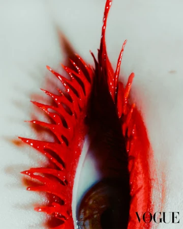 Macro beauty shot of a brown eye with dramatic, elongated bright red artistic eyelashes and makeup against a light grey background.