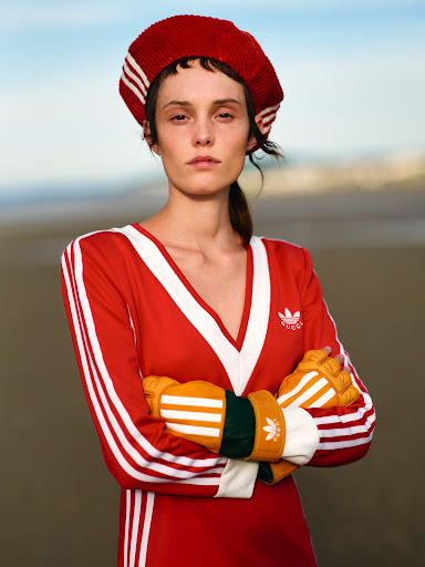 A fashion portrait of an adult female wearing a red and white Adidas tracksuit and matching hat, standing on a beach with crossed arms.