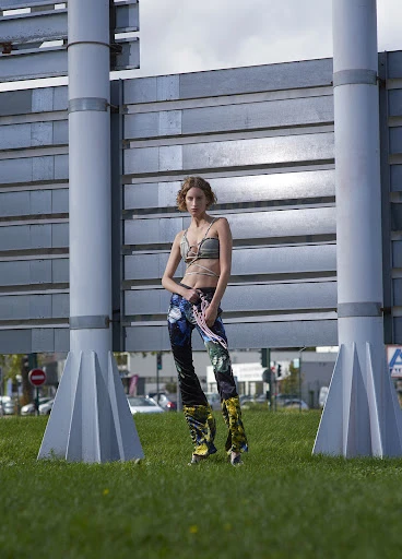 Young woman in stylish patterned pants and top poses confidently on roller skates near a metallic industrial structure on a grassy field.